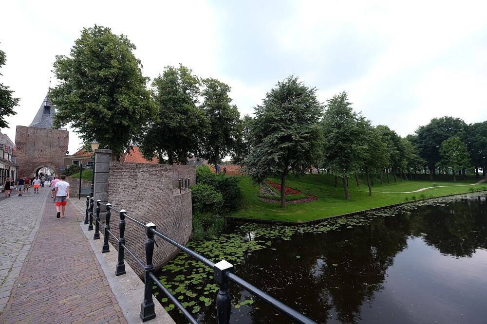 Bakstenen poorttoren met spits aan het einde van een gekasseide kade langs de vestinggracht van Elburg, met wandelaars en een groen bolwerk aan de overkant