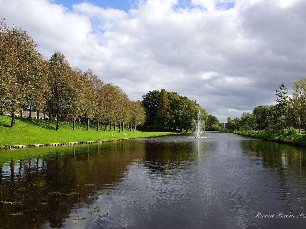 Brede vestinggracht met een fontein, aflopende grasoevers en een bomenrij met wandelpad bij Elburg